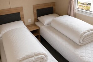 A hotel room with two single beds, a wooden headboard, a nightstand, and a window with curtains. - Serene Lodge Retreat in Clacton (Clacton-on-Sea)