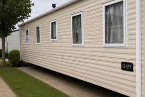 A row of mobile homes with a flat roof, white siding, and a small front yard with grass and a bush. - Serene Lodge Retreat in Clacton (Clacton-on-Sea)