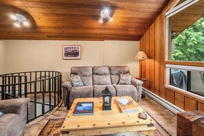 A cozy living room with a wooden ceiling, a large window, a sofa with decorative pillows, a coffee table with books and a lantern, and a crib. - Limber Pine Townhome and 2BDR Stay (Whitefish)