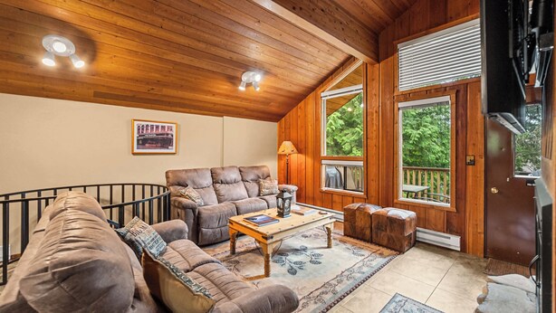 Ein gemütliches Wohnzimmer mit Holzbalkendecke, einer Sofagarnitur, einem Couchtisch und Blick ins Freie durch ein großes Fenster.