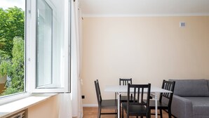 Um quarto pequeno e bem iluminado, com uma mesa de jantar e cadeiras, um sofá cinzento e uma janela com cortinas.
