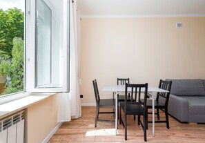 Ein kleiner, gut beleuchteter Raum mit einem Esstisch und Stühlen, einem grauen Sofa und einem Fenster mit Vorhängen.