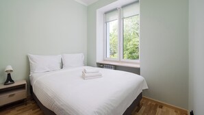 Quarto com cama, almofadas, mesa de cabeceira, luminária e janela com vista para árvores.