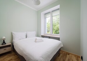 Ein Schlafzimmer mit Bett, Kissen, einem Nachttisch, einer Lampe und einem Fenster mit Blick auf Bäume.