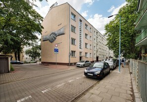 Ein Gebäude mit einer großen Wandmalerei von zwei sich streckenden Händen, einem Verbotsschild für Parkverbote und parkenden Autos entlang der Straße.