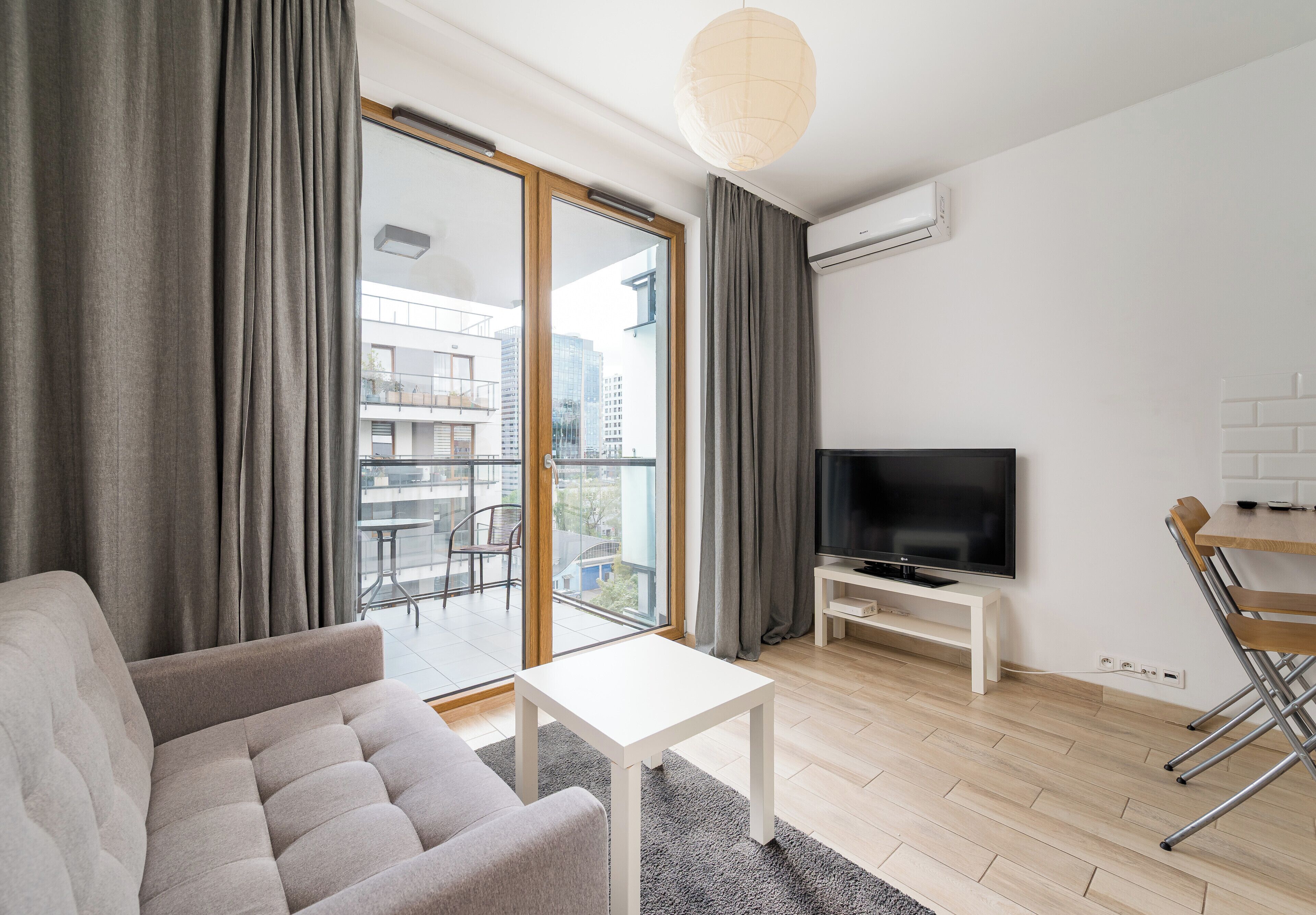 A modern living room with a sofa, a flat-screen TV, a small table, and a balcony with a view of buildings.