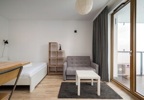 A modern living room with a bed, a grey sofa, a small white table, a wooden bookshelf, and a large window with curtains. - Wola Jaktorowska Urban Studio (Warszawa)