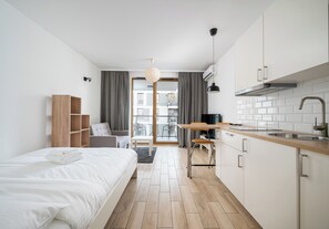 A modern, compact hotel room with a bed, a small kitchenette, a dining area, and a view of the city through the window. - Wola Jaktorowska Urban Studio (Warszawa)