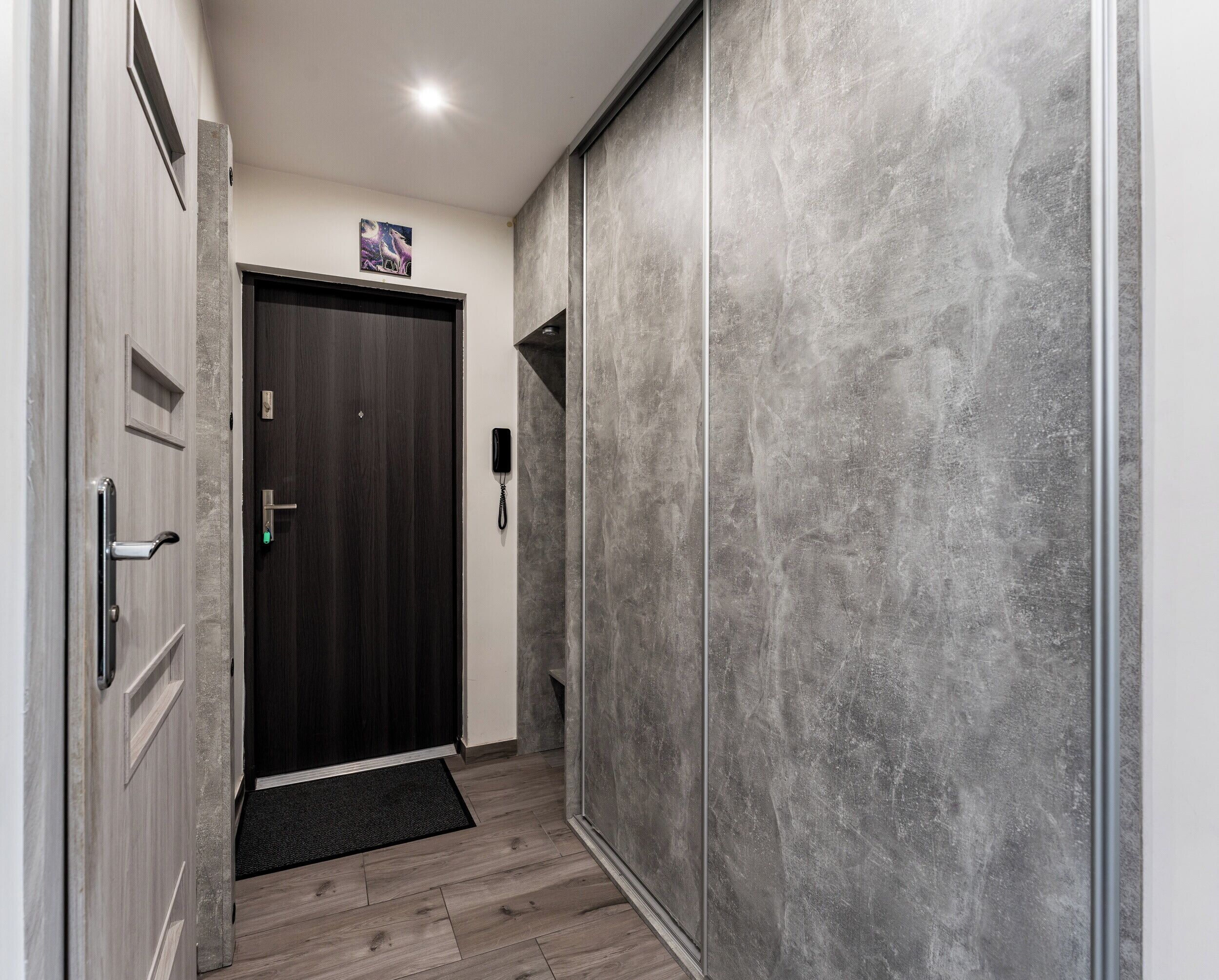 Un couloir avec une porte en bois sombre, un téléphone fixé au mur et un mur gris à texture.