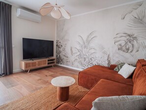 A modern living room with a flat-screen TV, a wooden entertainment unit, a brown sofa, a round coffee table, and a tropical leaf-patterned wall. - Penthouse, 200m from the beach w/ parking (Cubelles)