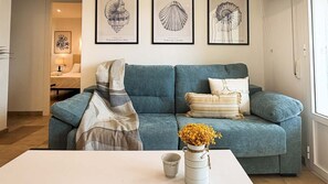 Ein Wohnzimmer mit einem petrolfarbenen Sofa, einem weißen Couchtisch und einer Vase mit gelben Blumen. Über dem Sofa hängen gerahmte Bilder an der Wand.