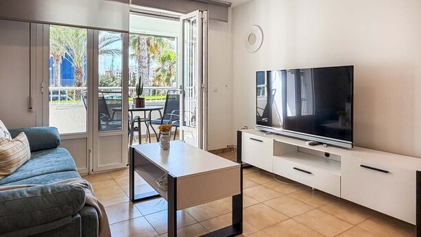 A modern living room with a sofa, a coffee table, a TV on a stand, and a sliding glass door leading to an outdoor area with a table and chairs. - Pink Las Olas Apartment (Punta Prima)