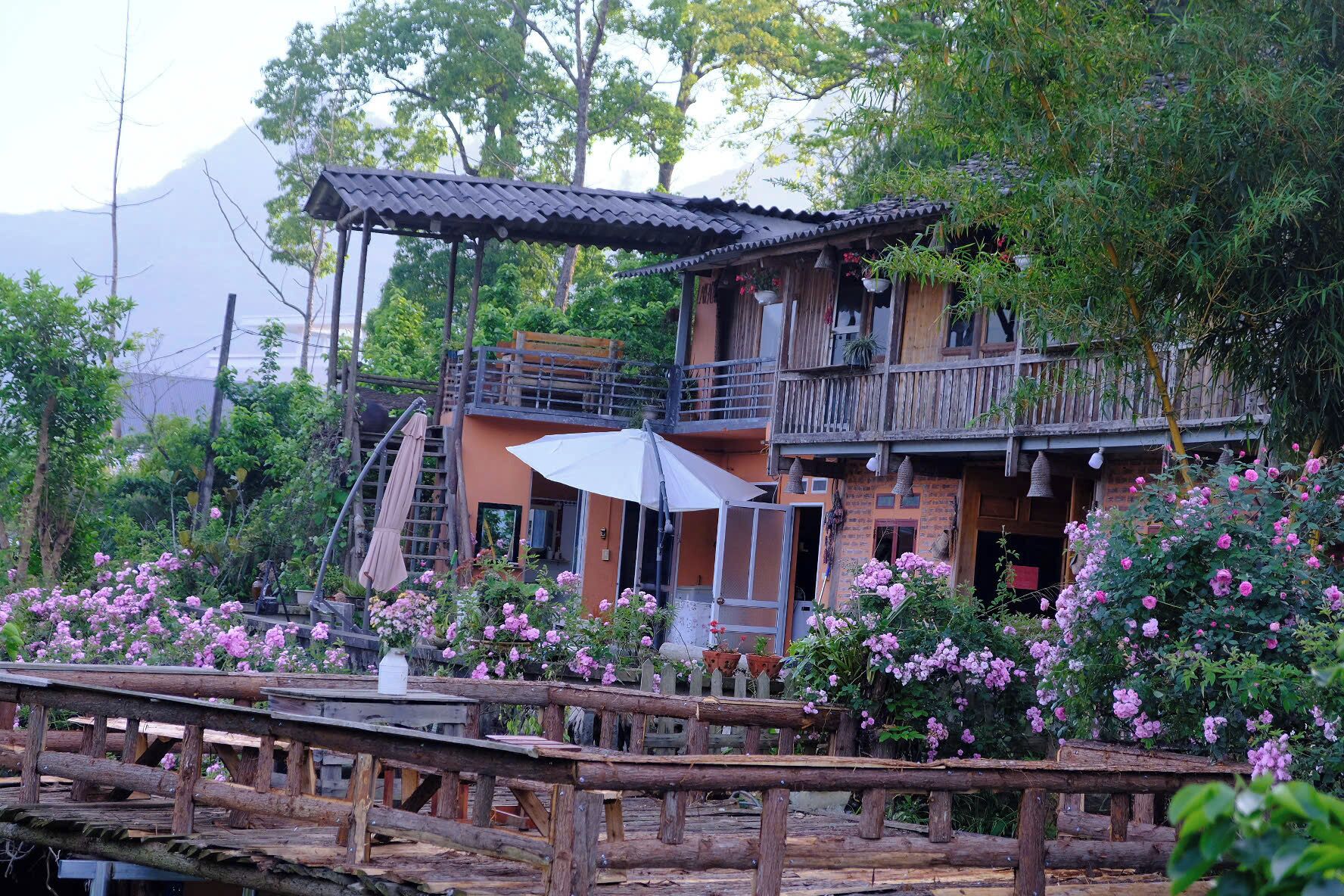 Une maison en bois au style rustique, dotée d’un balcon et entourée d’une végétation luxuriante et de fleurs roses.
