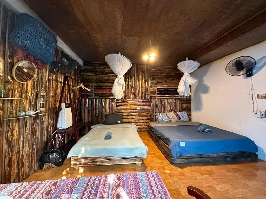 Quarto com duas camas, parede de madeira, tapete estampado e ventilador de teto.