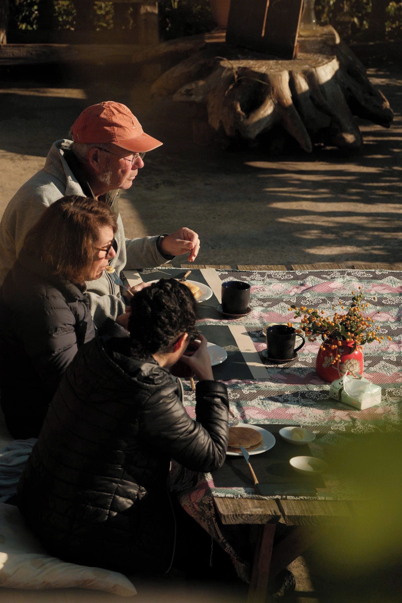 Un groupe de personnes est assis autour d’une table en bois, à l’extérieur, sur laquelle est posée une nappe à motifs, et elles savourent un repas.