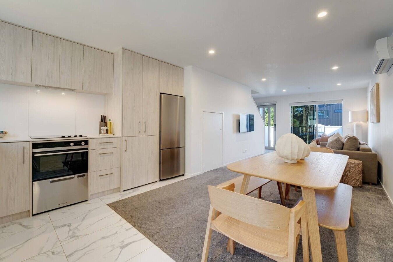 A modern kitchen with built-in appliances, a dining area with a wooden table and chairs, and a view of the outdoors through large windows.