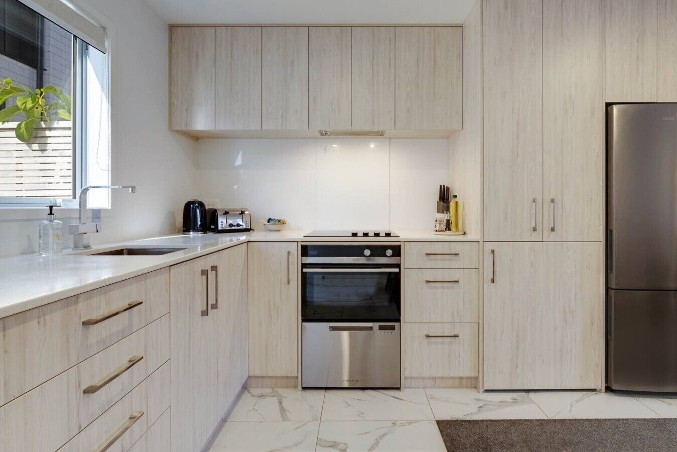 A modern kitchen with light wood cabinets, stainless steel appliances, and a marble floor.
