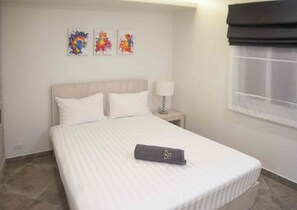 Ein Hotelzimmer mit einem Bett, weißer Bettwäsche, einem Nachttisch mit Lampe und drei farbenfrohen Bildern an der Wand.