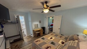 Ein Schlafzimmer mit einem Bett, einem Schreibtisch, einem Schminktisch mit Beleuchtung, einem Deckenventilator und einer Tür zu einem anderen Raum.
