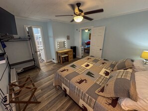 Ein Schlafzimmer mit einem Bett, einem Schreibtisch, einem Schminktisch mit Beleuchtung, einem Deckenventilator und einer Tür zu einem anderen Raum.
