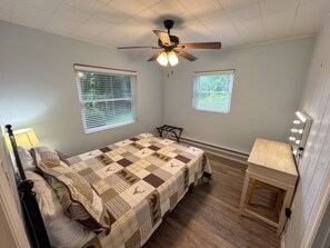 Ein Schlafzimmer mit einem Bett, einem Deckenventilator, zwei Fenstern, einem Nachttisch und einer Lampe.