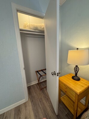 Ein Zimmer mit einem Nachttisch aus Holz, einer Lampe, einem Holzhocker und einem Kleiderschrank mit Kleiderstange.