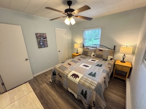 A bedroom with a bed, two bedside tables with lamps, a ceiling fan, a window with blinds, and a door. - The Honey Bear Retreat nestled in the heart of Maggie Valley (Maggie Valley)