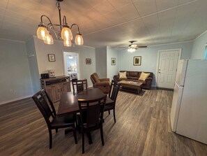 Ein Essbereich mit Holztisch und -stühlen, eine Küche mit Kühlschrank und ein Wohnzimmer mit Sofa und Deckenventilator.