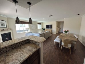 A modern kitchen with a marble countertop, a dining area with a wooden table and chairs, and a living area with a sofa and television. - Steps to Mission Bay! 2BR Townhome w/Roof Deck, Sea World Drone Show & Bay Views (San Diego)