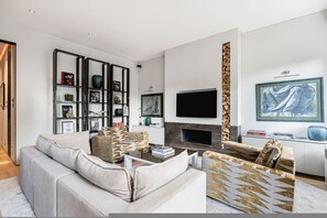 Ein stilvolles Wohnzimmer mit einer Couch, Sesseln, einem Couchtisch, einem Kamin, einem Fernseher und einem Bücherregal.