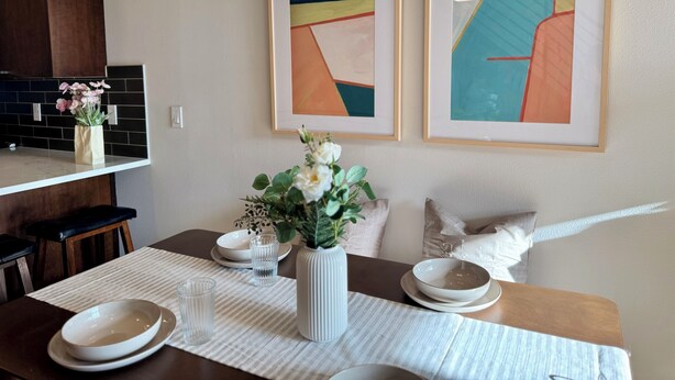 Uma área de jantar com uma mesa posta para dois, um vaso com flores e dois quadros abstratos emoldurados na parede.
