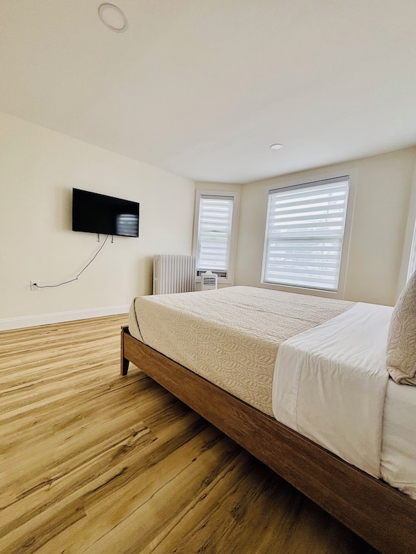 Un dormitorio con suelo de madera, una cama con ropa blanca, un televisor montado y dos ventanas con persianas.
