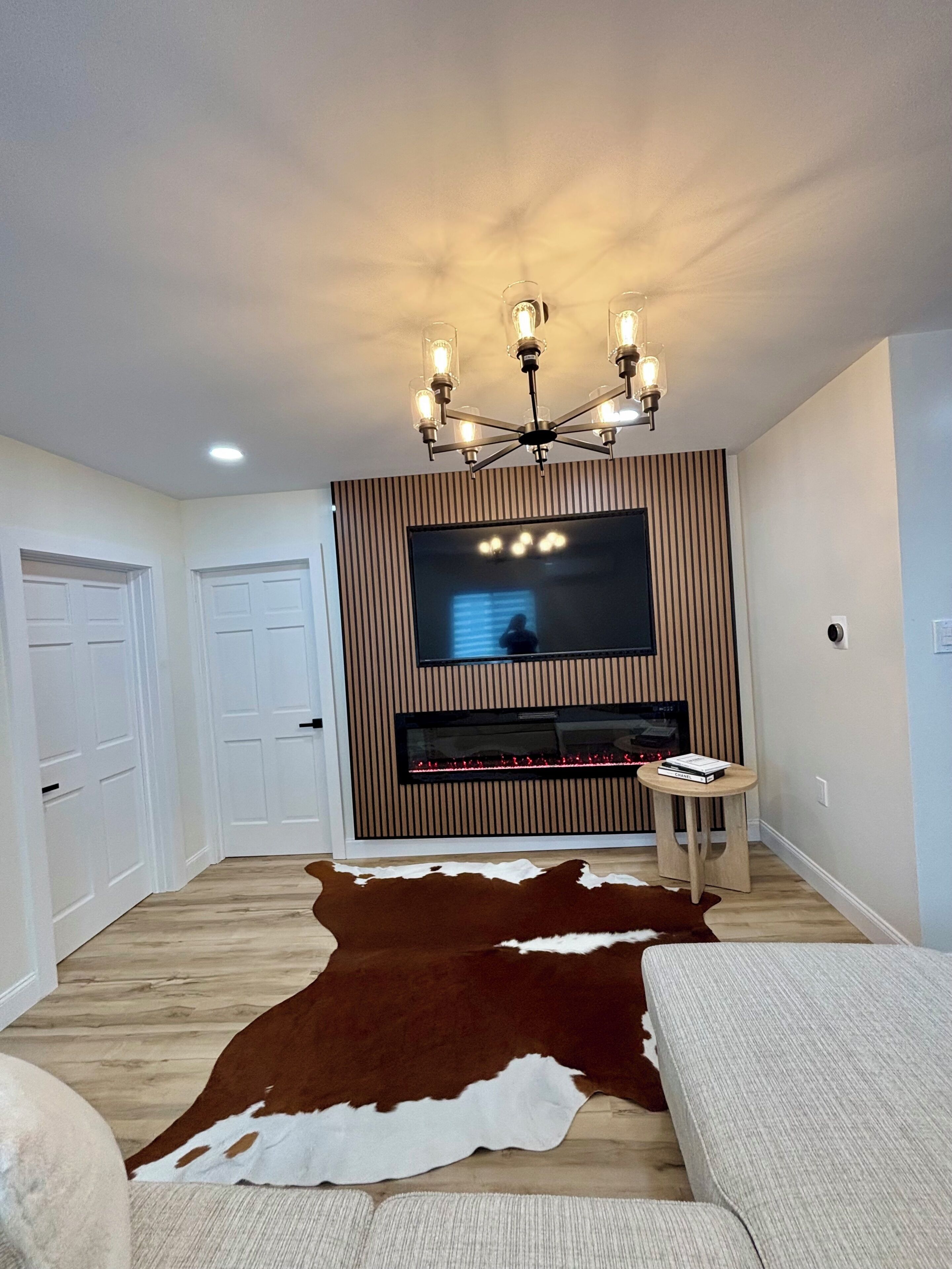 Un salon doté d’un téléviseur à écran plat fixé sur un mur recouvert de panneaux en bois, d’un lustre, d’une cheminée et d’un tapis en peau de vache.