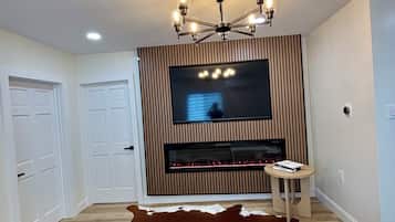 Un salon doté d’un téléviseur à écran plat fixé sur un mur recouvert de panneaux en bois, d’un lustre, d’une cheminée et d’un tapis en peau de vache.