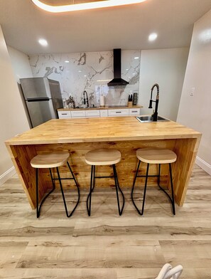 A kitchen with a large wooden island, three stools, a stainless steel refrigerator, and a marble backsplash.