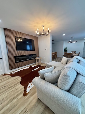 Una sala de estar moderna con un televisor de pantalla plana grande, un sofá, una araña y un comedor al fondo.