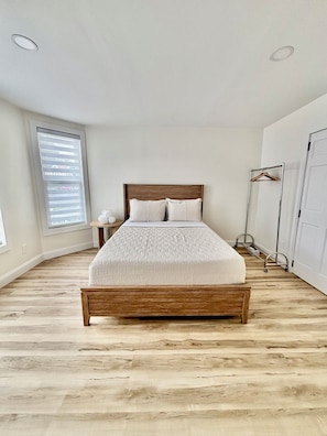 A bedroom with a wooden bed, a bedside table, a hanging light fixture, a wardrobe, and a window with blinds.