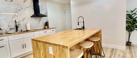 Moderni keittiö, jossa on puinen keittiösaareke, baarituolit, valkoiset kaapit ja musta liesituuletin.