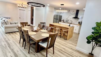 A modern kitchen with a central island, a dining area with a wooden table and chairs, and a living area with a sofa and a plant.