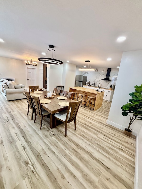 A modern kitchen with a central island, a dining area with a wooden table and chairs, and a living area with a sofa and a plant.