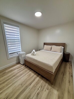 A bedroom with a wooden bed, a window with blinds, a standing heater, and a round wooden stool.
