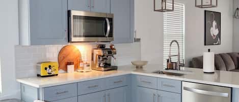 A modern kitchen with blue cabinets, a stainless steel microwave, a coffee machine, and a dishwasher.