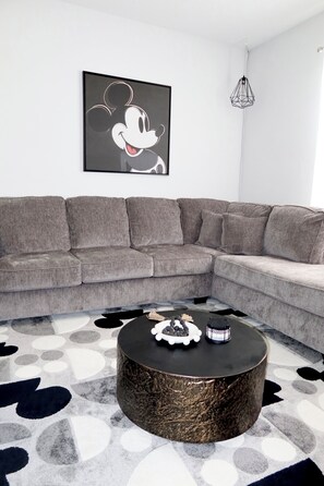 Ein modernes Wohnzimmer mit einem grauen Ecksofa, einem runden Couchtisch mit Dekoration und einem Wandbild mit Mickey Mouse.