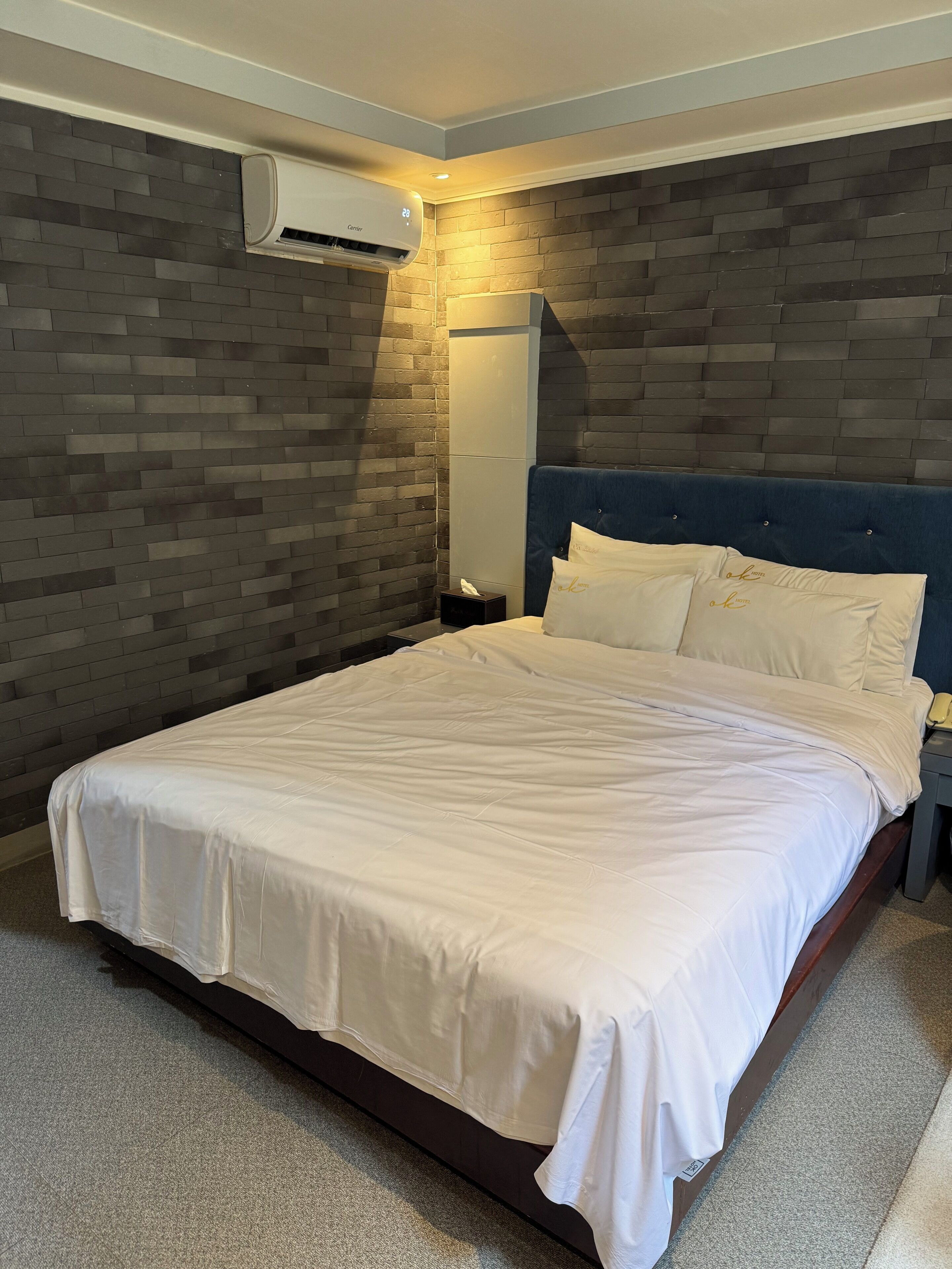 A hotel room with a large bed, a bedside table with a lamp, a wall-mounted air conditioner, and a brick-patterned wall.