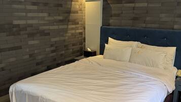 A hotel room with a large bed, a bedside table with a lamp, a wall-mounted air conditioner, and a brick-patterned wall.