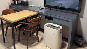 A hotel room with a bed, a desk with a computer, a chair, a TV, a mirror, and a waste bin.