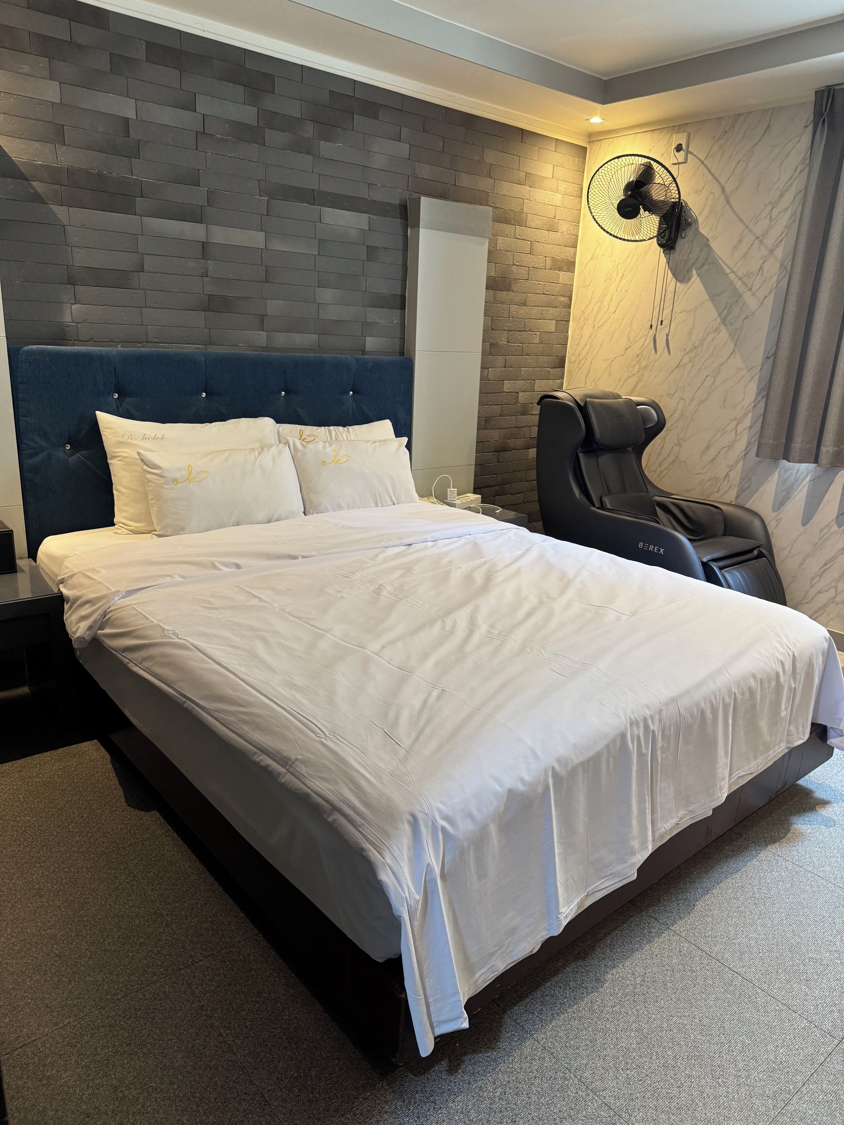 A hotel room with a large bed, a massage chair, a wall-mounted fan, and a brick-patterned wall.