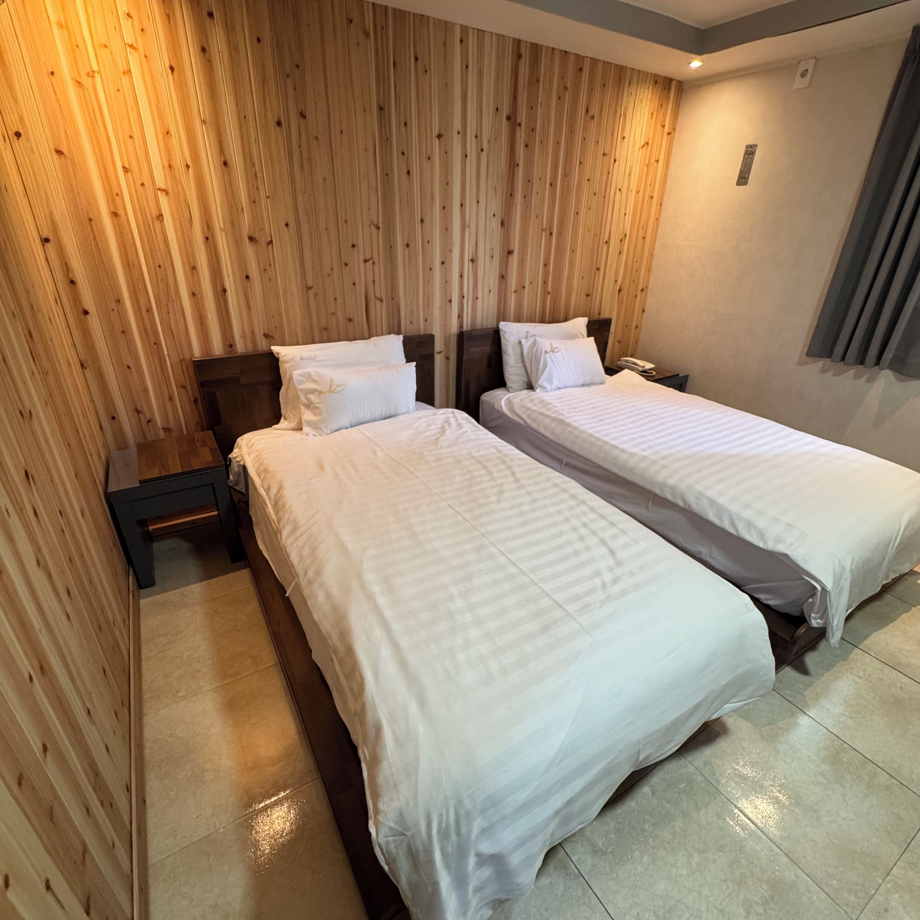 A hotel room with two beds, wooden panel walls, a nightstand with a phone, and a window with curtains.
