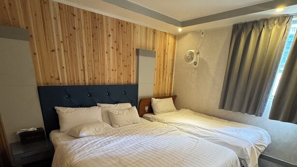 A hotel room with two beds, a wooden panel wall, a ceiling fan, and a window with curtains.