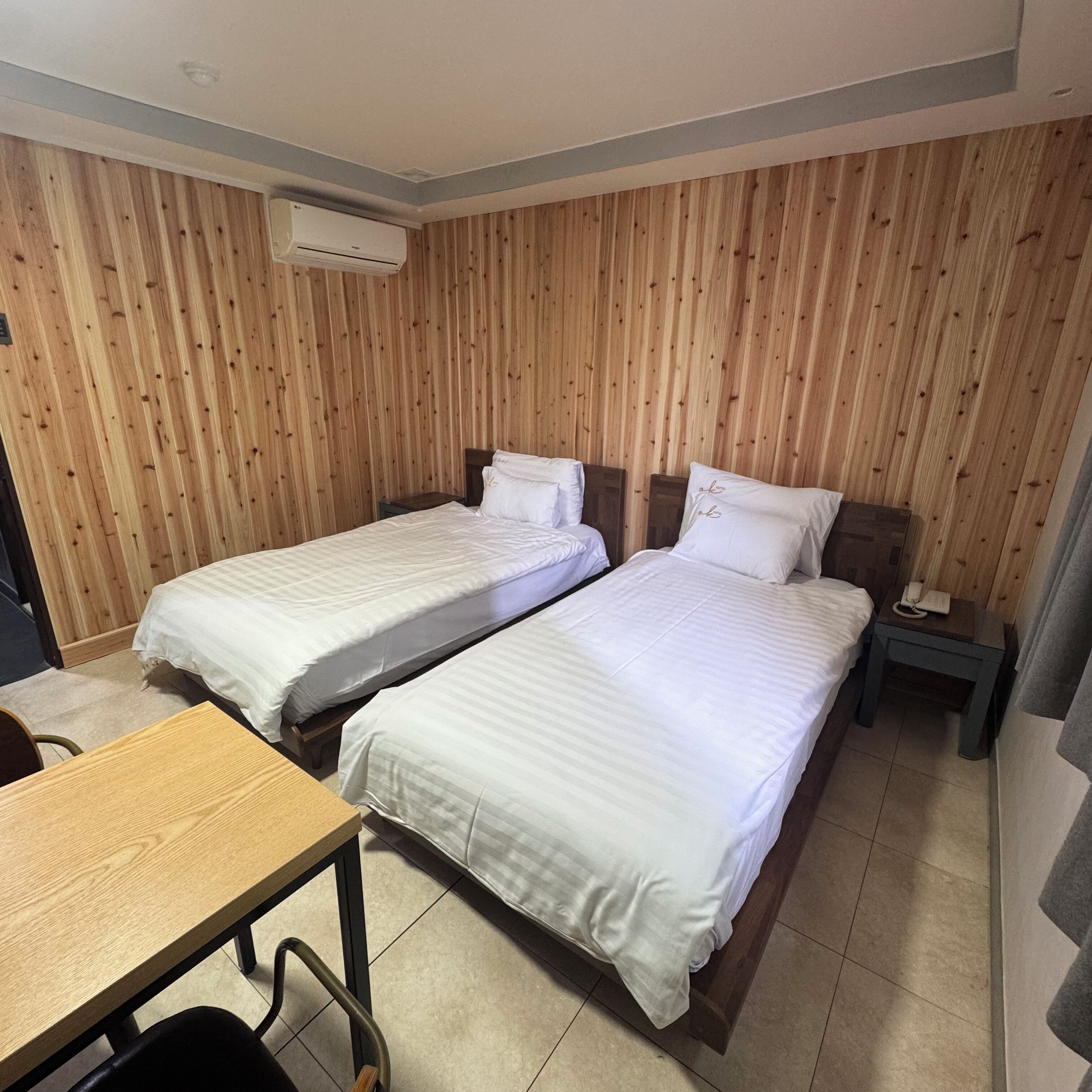 Una habitación de hotel con dos camas individuales, paneles de madera en la pared, una mesita pequeña y una mesita de noche con un teléfono.